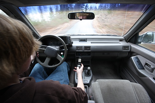 A young teen texting on his flip-phone as he drives down the road. Mirror shows that his eyes are on the phone, not on the road. Blurred motion out the windshied and speed on the speedometer show that he's really driving. Phone screen says "lol".